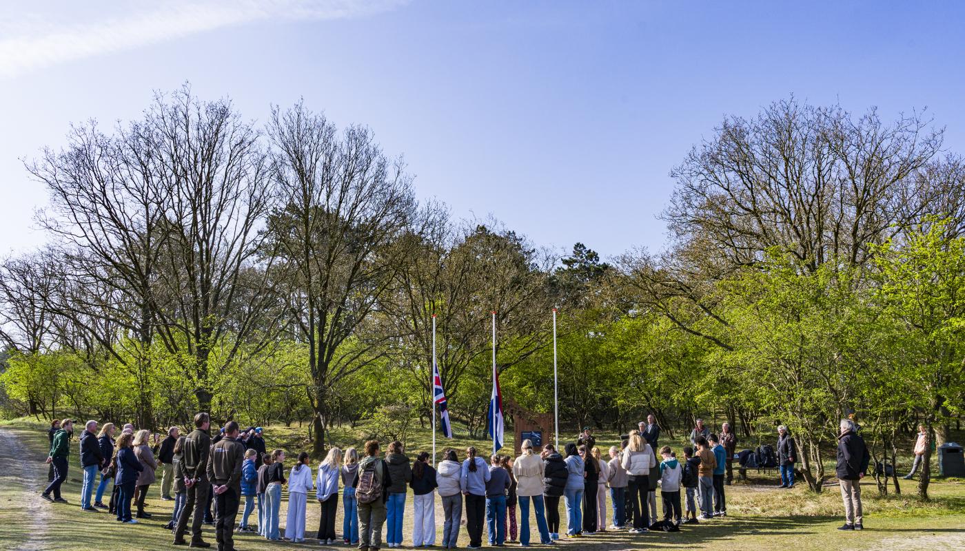 Herdenkingsceremonie bij de AWD, waarbij meerdere mensen aandachtig toekijken terwijl vlaggen halfstok hangen.